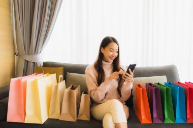 Portre güzel genç Asyalı kadın dizüstü bilgisayar ya da akıllı cep telefonu kullanarak oturma odasındaki alışveriş çantasının etrafındaki kanepede alışveriş yapıyor.