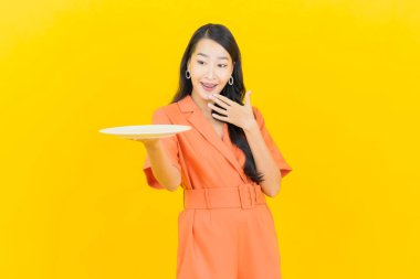 Asyalı güzel bir kadının portresi. Sarı arka planda boş tabak tabağıyla gülümsüyor.