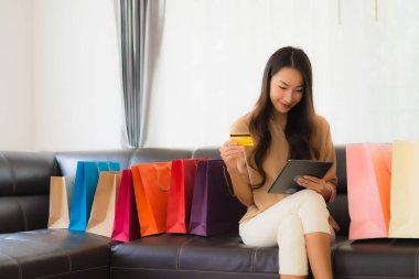 Güzel Asyalı bir kadının portresi. İnternetten alışveriş yapıyor. Kredi kartı ve akıllı telefonuyla oturma odasındaki alışveriş çantasının etrafında.
