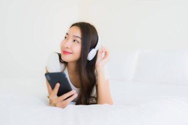 Asyalı güzel bir kadının portresi mutlu bir gülümseme akıllı cep telefonu ve kulaklıkla yatak odasının içinde müzik dinlemek için.