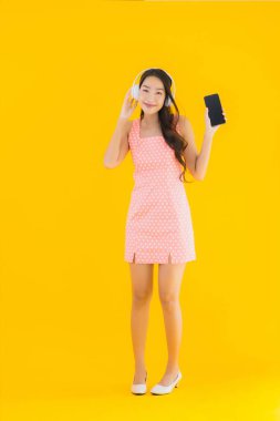 Güzel bir Asyalı kadın portresi. Sarı izole edilmiş arka planda akıllı cep telefonu ve kulaklıkla müzik dinliyor.
