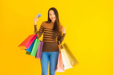 Güzel bir Asyalı kadının portresi sarı izole edilmiş arka planda renkli alışveriş torbalarıyla alışveriş yapmak için kredi kartı kullanıyor.