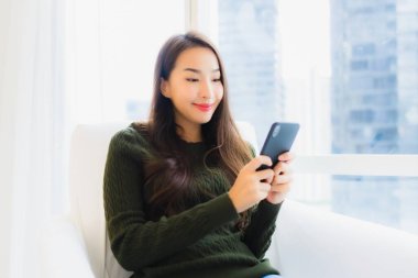 Güzel bir Asyalı kadın oturma odasındaki kanepede akıllı cep telefonu kullanıyor.