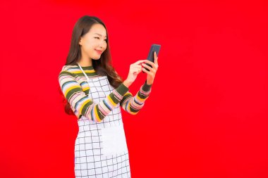 Güzel bir Asyalı kadın portresi kırmızı izole edilmiş arka planda akıllı cep telefonuyla önlük giyiyor.