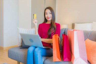 Güzel bir Asyalı kadın portresi. Kanepede çantalı online alışveriş için bilgisayarla kredi kartı kullanıyor.