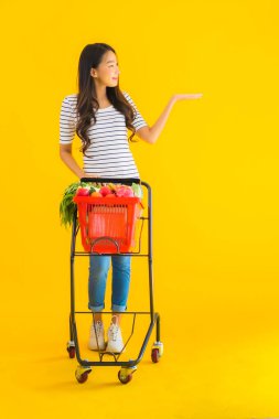 Güzel genç Asyalı kadın süpermarketten alışveriş yapıyor ve sarı izole arka planda el arabası var.