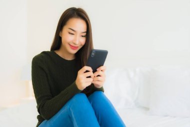 Güzel bir Asyalı kadın portresi. Yatak odasında beyaz yastık battaniyesi ve yatağında akıllı cep telefonu kullanıyor.