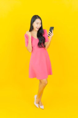 Asyalı güzel bir kadının portresi sarı arka planda akıllı cep telefonuyla gülümsüyor.