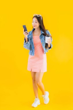 Kahve fincanı ve sarı izole edilmiş arka planda akıllı cep telefonlu güzel Asyalı kadın portresi.