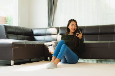 Güzel bir Asyalı kadın portresi oturma odasının iç tarafında kanepeli bir kahve fincanı ve akıllı cep telefonu kullanıyor.