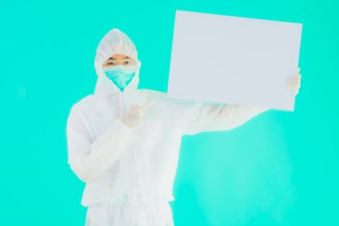 Güzel, genç Asyalı doktor kadın koronavirüsten korunmak için ppe ya da kişisel koruyucu ekipman takıyor ya da mavi izole arka planda beyaz kartı olan 19. bölgede yaşıyor.