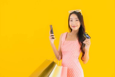 Renkli alışveriş çantalı güzel Asyalı kadın akıllı cep telefonu ve sarı izole edilmiş arka planda kredi kartı.