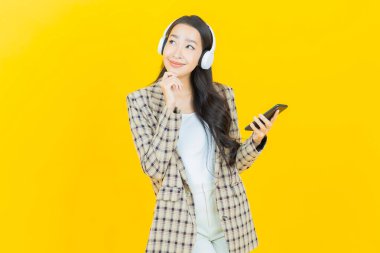 Sarı arka planda müzik dinlemek için kulaklığı ve akıllı telefonu olan güzel Asyalı kadın portresi.
