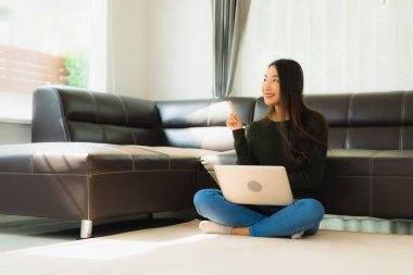 Güzel bir Asyalı kadın portresi. Oturma odasında kanepesi olan bir dizüstü bilgisayar kullanıyor.
