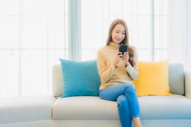 Oturma odasındaki kanepede akıllı cep telefonlu güzel Asyalı kadın portresi.