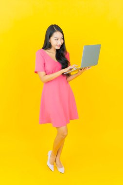 Asyalı güzel bir kadının portresi. Sarı izole edilmiş arka planda dizüstü bilgisayarla gülümsüyor.