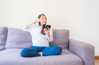 Güzel bir Asyalı kadın oturma odasında müzik dinlemek için akıllı cep telefonu kulaklığı kullanıyor.
