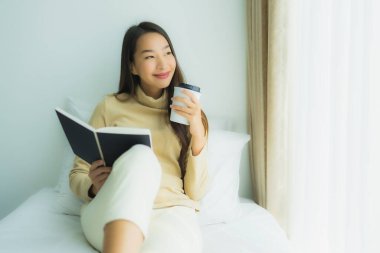 Genç Asyalı kadın, elinde kahve fincanıyla yatak odasında kitap okuyor.
