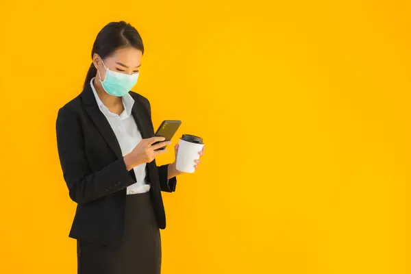 Portrait beautiful young business asian woman wear mask use mobile phone with coffee for covid19 and coronavirus concept on yellow isolated background