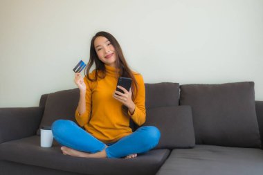 Genç Asyalı kadının oturma odasında online alışveriş yapmak için akıllı cep telefonu ve kredi kartı olan dizüstü bilgisayarı kullandığı portre.