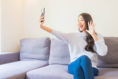 Güzel bir Asyalı kadın oturma odasındaki koltukta akıllı cep telefonu kullanıyor.
