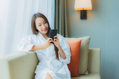Güzel bir Asyalı kadın oturma odasının iç tarafındaki koltukta akıllı cep telefonu kullanıyor.