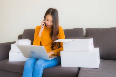 Asyalı genç bir kadının portresi, oturma odasındaki koltukta paket kutusuyla çalışmak için dizüstü bilgisayar kullanıyor.