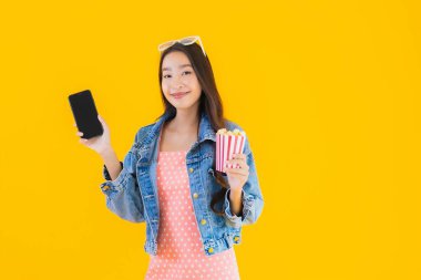 Güzel bir Asyalı kadın portresi telefon patlamış mısırı ve sarı izole edilmiş bir arka planda film izlemekten memnun.