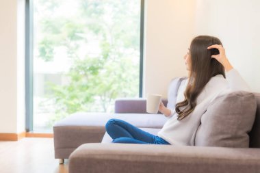 Oturma odasındaki kanepede kahve fincanı taşıyan güzel Asyalı kadın portresi.