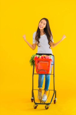 Güzel genç Asyalı kadın süpermarketten alışveriş yapıyor ve sarı izole arka planda el arabası var.