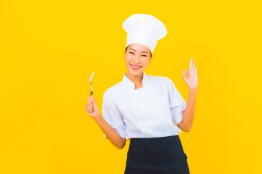 Güzel bir portre genç Asyalı kadın aşçı ya da aşçı üniforması giyiyor sarı izole arka planda çatal ve kaşıkla.