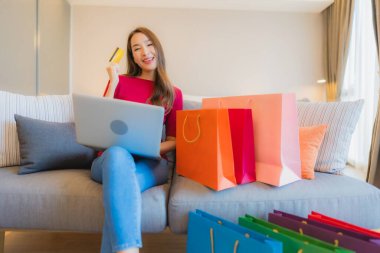 Güzel bir Asyalı kadın portresi. Kanepede çantalı online alışveriş için bilgisayarla kredi kartı kullanıyor.