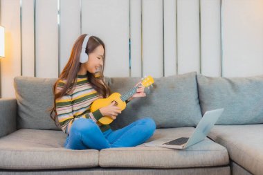 Güzel bir Asyalı kadın oturma odasındaki kanepede ukulele çalıyor.