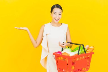 Asyalı güzel bir kadının portresi. Sarı arka planda süpermarketten alınmış alışveriş sepetiyle gülümsüyor.