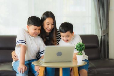 İki oğlu olan güzel Asyalı kadın anne oturma odasındaki kanepede dizüstü bilgisayar ya da bilgisayar kullanıyor.