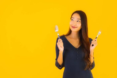 Güzel, genç Asyalı bir kadının portresi. Kaşığı ve çatalı ile sarı izole edilmiş arka planda yemeye hazır.