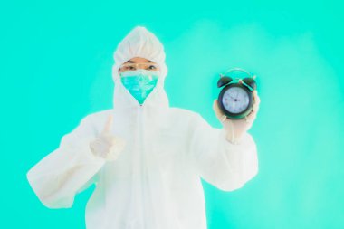 Güzel bir portre genç Asyalı doktor. Özel süit ve maske takıyor. Mavi arka planda saati ya da alarmı gösteriyor.