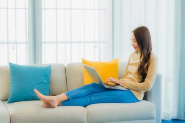 Asyalı güzel bir kadının portresi oturma odasının iç dekorasyonunda dizüstü bilgisayar kullanıyor.