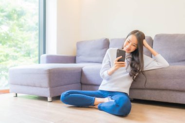 Güzel bir Asyalı kadın oturma odasındaki koltukta akıllı cep telefonu kullanıyor.