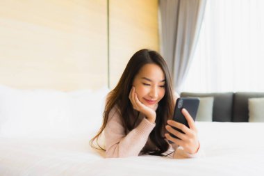 Yatak odasındaki yatağın üstünde kahve fincanı ve cep telefonu olan güzel Asyalı kadın portresi.