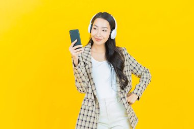 Sarı arka planda müzik dinlemek için kulaklığı ve akıllı telefonu olan güzel Asyalı kadın portresi.