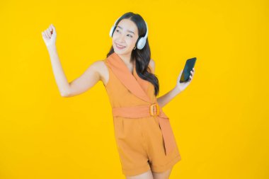 Sarı arka planda müzik dinlemek için kulaklığı ve akıllı telefonu olan güzel Asyalı kadın portresi.