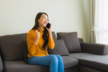 Genç Asyalı kadının oturma odasında yastıklı akıllı cep telefonu kullandığı portre.