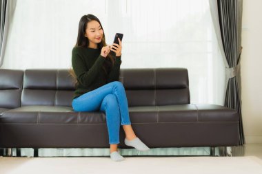Güzel bir Asyalı kadın oturma odasındaki koltukta akıllı cep telefonu ya da cep telefonu kullanıyor.