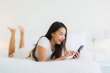 Güzel bir Asyalı kadın portresi. Yatak odasında beyaz yastık battaniyesi ve yatağında akıllı cep telefonu kullanıyor.