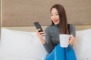 Güzel bir Asyalı kadın yatak odasının iç kısmında, içinde kahve fincanı olan akıllı bir cep telefonu kullanıyor.