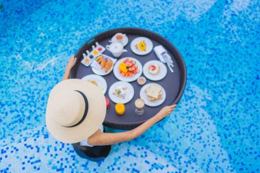 Asyalı güzel bir kadının portresi yüzme havuzunda yüzen kahvaltı tepsisiyle mutlu bir gülümseme