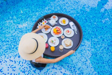 Asyalı güzel bir kadının portresi yüzme havuzunda yüzen kahvaltı tepsisiyle mutlu bir gülümseme