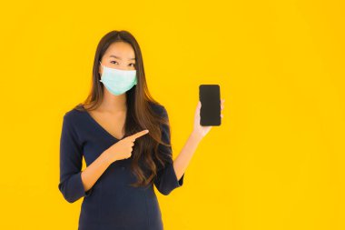 Maskeli ve akıllı cep telefonlu güzel bir Asyalı kadın portresi veya izole edilmiş sarı arka planda cep telefonu.