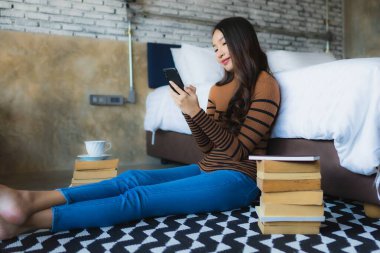 Genç Asyalı kadın yatak odasının içinde kitap ve kahve fincanı ile akıllı cep telefonu kullanıyor.